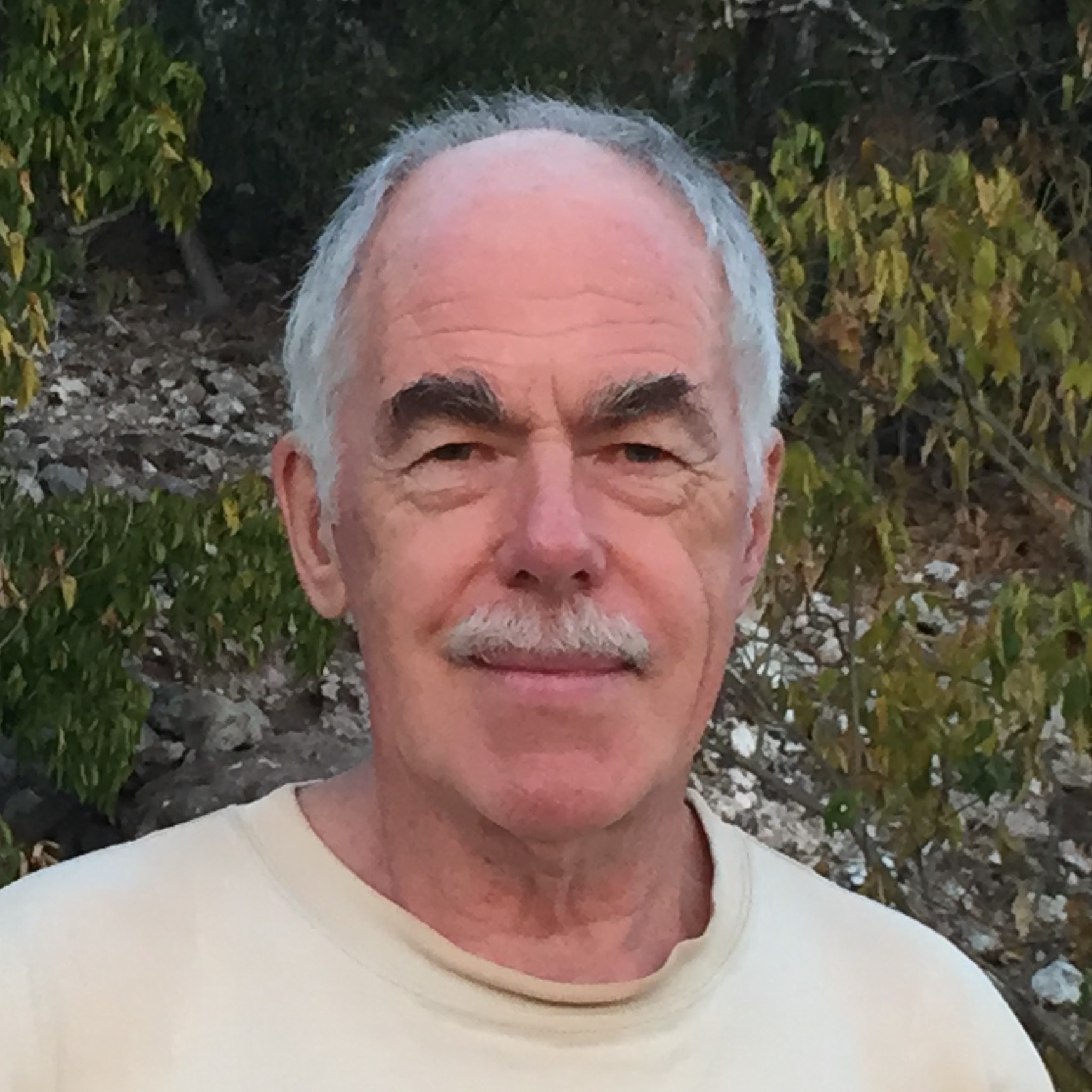 Hugh Drummond with short gray hair and a moustache in a tan t-shirt in front of some trees.